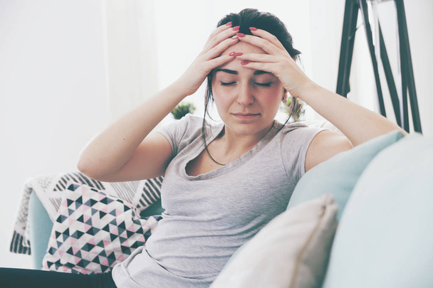 Young worried woman with headache while sitting on comfortable sofa - Φωτογραφία, εικόνα