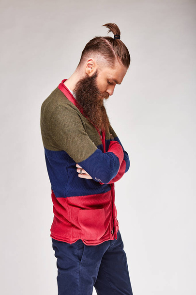 Young bearded man with trendy hair posing in studio in trendy modern colored sweater with red, dark blue and olive stripes and blue chinos - Foto, immagini
