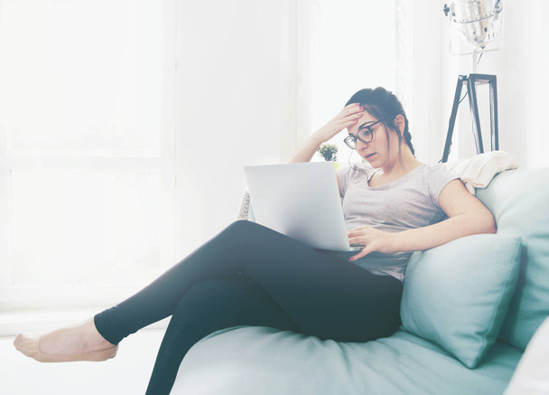 Mujer preocupada joven usando el ordenador portátil mientras está sentado en un cómodo sofá
 - Foto, imagen