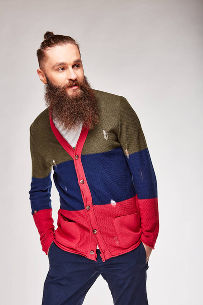 Young bearded man posing in studio in trendy modern colored sweater with red, dark blue and olive stripes and blue chinos - Φωτογραφία, εικόνα