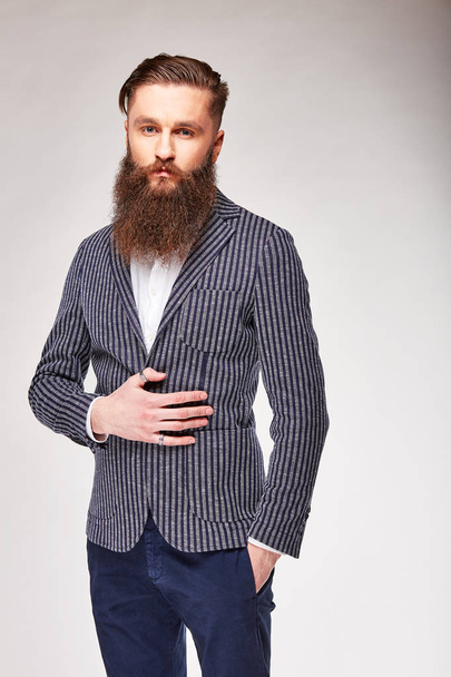 Young bearded man posing in studio in modern dark blue blazer with white stripes, white shirt and blue pants - Photo, image