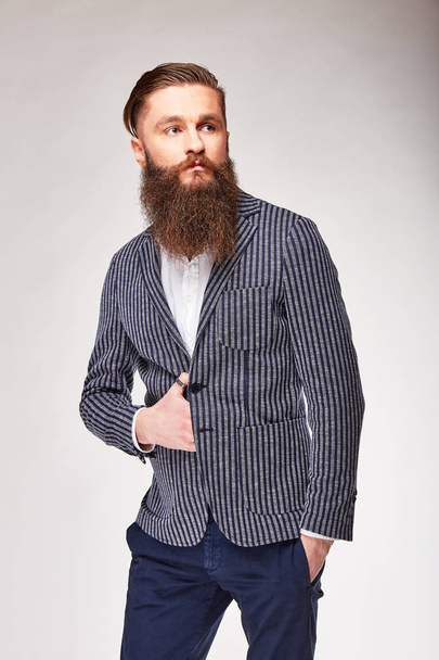 Young bearded man posing in studio in modern dark blue blazer with white stripes, white shirt and blue pants - 写真・画像