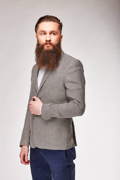 Young bearded man posing in studio in grey trendy blazer, white shirt and blue pants - Photo, image