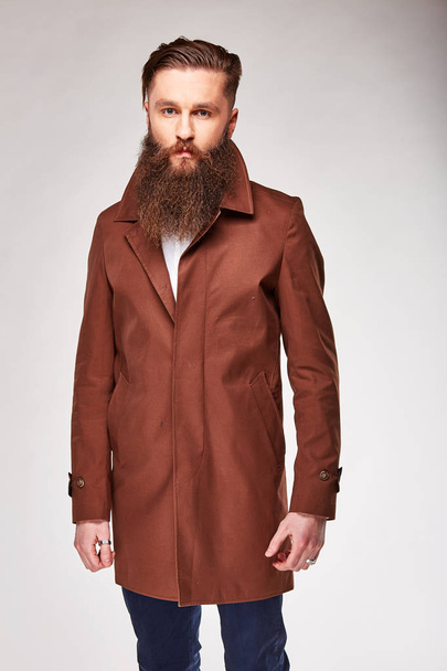 Young bearded man posing in studio in brown long jacket and blue pants - Фото, изображение