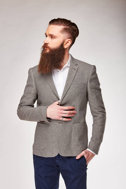 Young bearded man standing in studio in grey trendy blazer, white shirt and blue pants - Foto, immagini
