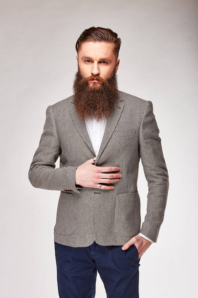 Young bearded man standing in studio in grey trendy blazer, white shirt and blue pants - 写真・画像