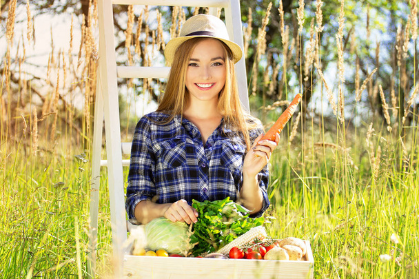 Pretty and young girl in hat, with wooden box full of vegetables and keeping carrot in her hand. Summer harvest. - Фото, изображение