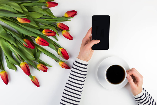 Manos de mujer con taza de café, teléfono móvil y hermoso ramo de flores en la mesa de oficina blanca
 - Foto, imagen