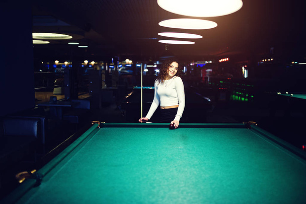 Young curly girl posed near billiard table. Sexy model at black  - 写真・画像