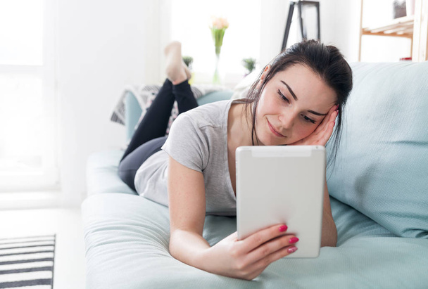 Young woman using pc tablet while lying on comfortable sofa - Foto, Imagem
