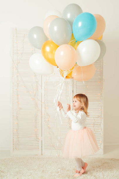 Счастливая маленькая девочка с тортом на день рождения и красочными шариками в комнате
. - Фото, изображение