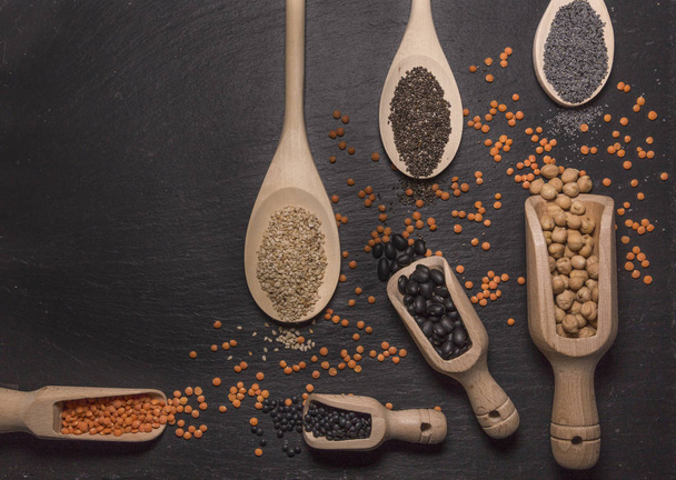 Sesame, chia and poppy seeds, red and black lentils, chickpeas and black beans with wooden spoons on black background - Foto, Bild