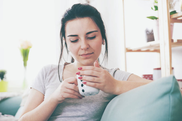 Young relaxed woman drinking coffee while sitting on comfortable sofa - Фото, изображение