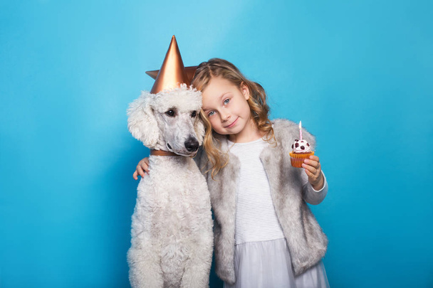 Little beautiful girl with dog celebrate birthday. Friendship. Love. Cake with candle. Studio portrait over blue background - Photo, Image