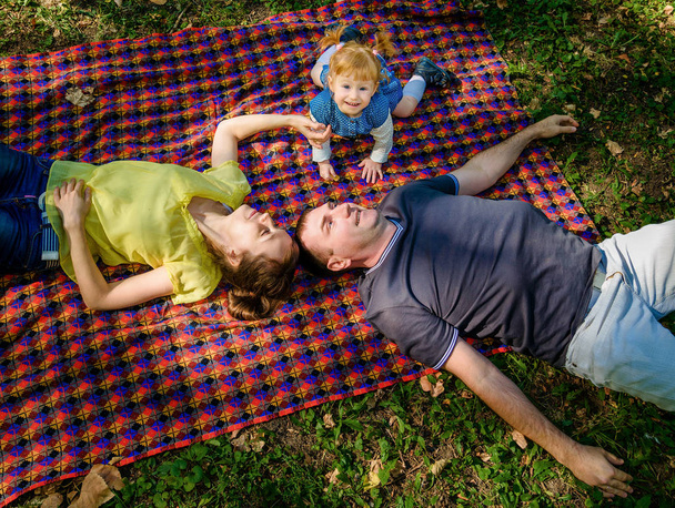 top view of young happy family of three persons. Father, mother and daughter lying on plaid and having fun in the park - Fotoğraf, Görsel