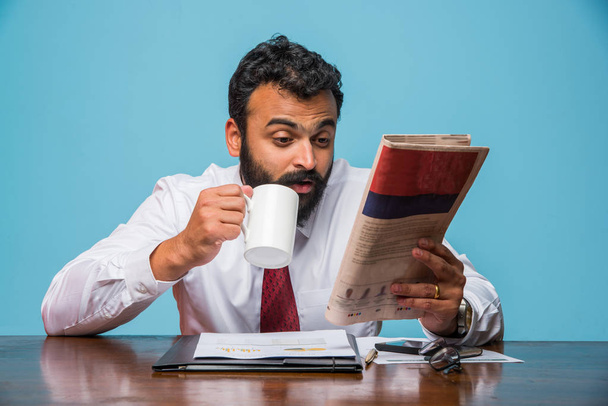 Foto s zvláštní efekt zobrazující indické mladý podnikatel čtenářský deník zároveň má kávy v kanceláři, asijský podnikatel si vychutnat kávu při čtení zprávy publikace - Fotografie, Obrázek
