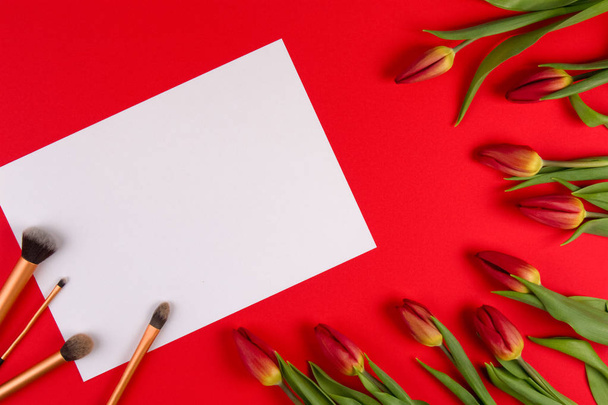 Componen los pinceles sobre el papel blanco blanco y las flores primaverales los tulipanes sobre el fondo rojo. Vista superior
 - Foto, imagen