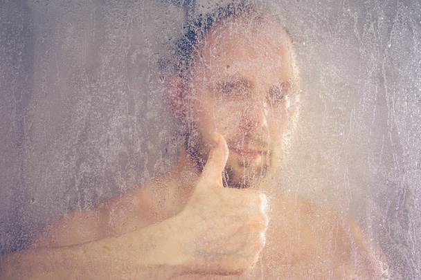 man taking a shower standing under flowing water and shows like sign in shower cabin behind transparent misted glass door in the bathroom - Photo, Image