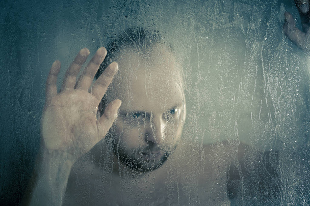 stressed man taking a shower standing under flowing water and holding his head in shower cabin behind transparent misted glass door in the bathroom - Foto, Imagem