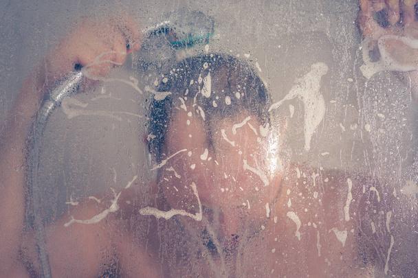 stressed man taking a shower standing under flowing water and holding his head in shower cabin behind transparent misted glass door in the bathroom - Photo, image