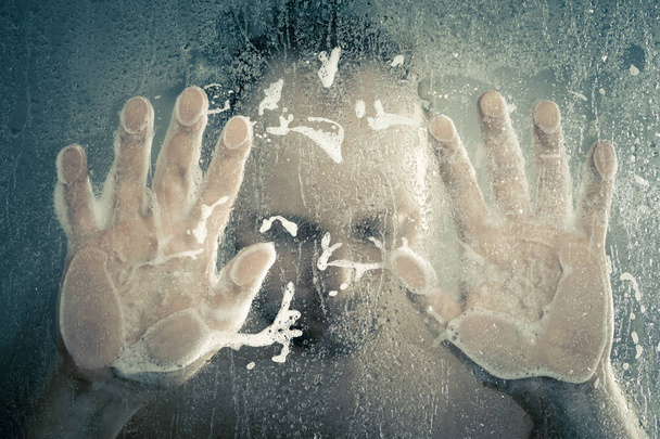 stressed man taking a shower standing under flowing water and holding his head in shower cabin behind transparent misted glass door in the bathroom - 写真・画像