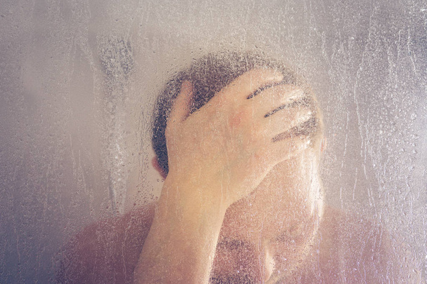 水が流れると浴室の透明な曇ったガラスのドアの後ろにシャワーの小屋で彼の頭を保持下に立ってシャワーを浴びて男を強調しました。 - 写真・画像