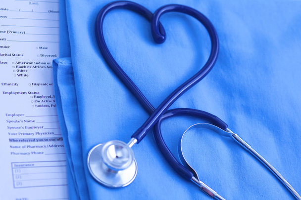 Ιατρικό στηθοσκόπιο στριμμένο σε σχήμα καρδιάς, που βρίσκεται στη λίστα του ιατρικού ιστορικού των ασθενών και ομοιόμορφη closeup μπλε γιατρό. Ιατρική βοήθεια ή ασφαλιστική έννοια. Καρδιολογική Φροντίδα, υγεία, προστασία και πρόληψη - Φωτογραφία, εικόνα
