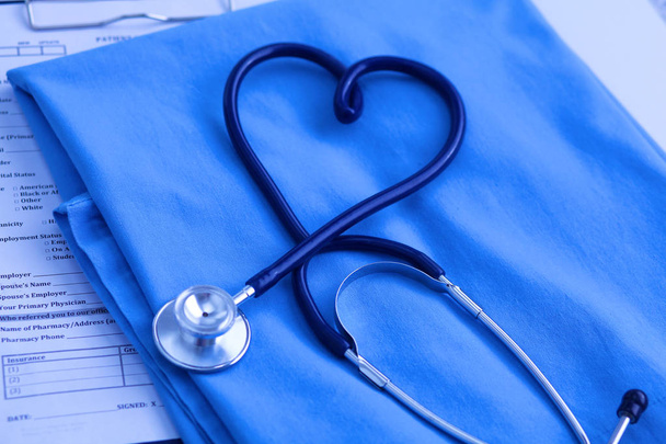 Ιατρικό στηθοσκόπιο στριμμένο σε σχήμα καρδιάς, που βρίσκεται στη λίστα του ιατρικού ιστορικού των ασθενών και ομοιόμορφη closeup μπλε γιατρό. Ιατρική βοήθεια ή ασφαλιστική έννοια. Καρδιολογική Φροντίδα, υγεία, προστασία και πρόληψη - Φωτογραφία, εικόνα