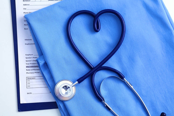 medizinisches Stethoskop in Herzform verdreht liegend auf Krankenakte und blauer Arztuniform Nahaufnahme. Medizinische Hilfe oder Versicherungskonzept. kardiologische Versorgung, Gesundheit, Schutz und Prävention - Foto, Bild