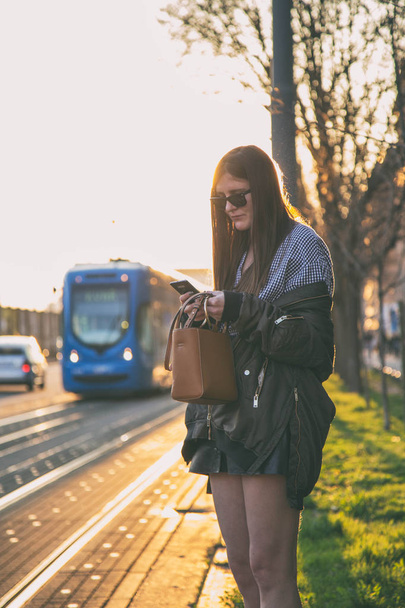 αστική νεαρή γυναίκα χρησιμοποιώντας smartphone της στο δρόμο για το ηλιοβασίλεμα της πόλης. Fashion blogger γραπτά μηνύματα στο τηλέφωνό της. κομψή γυναίκα χρησιμοποιώντας το τηλέφωνό της για τα κοινωνικά δίκτυα. online αγορές έννοια. - Φωτογραφία, εικόνα