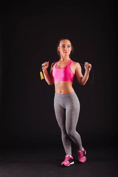 Young girl with a skipping rope in hands in her hands on a black background. The woman in pink top and gray leggings - Photo, Image