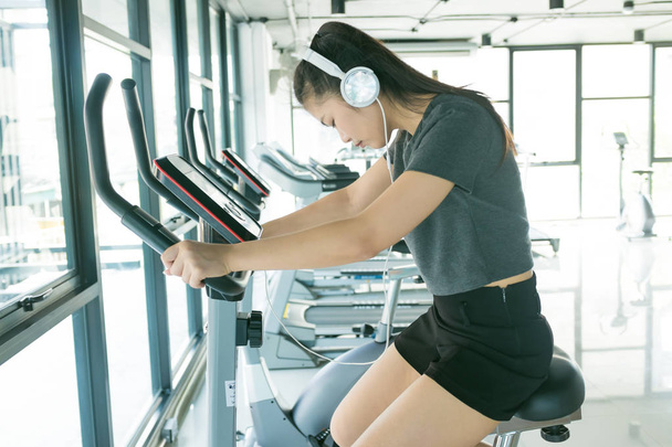 Fitness women cycling on bike and listening music in fitness gym - Photo, Image