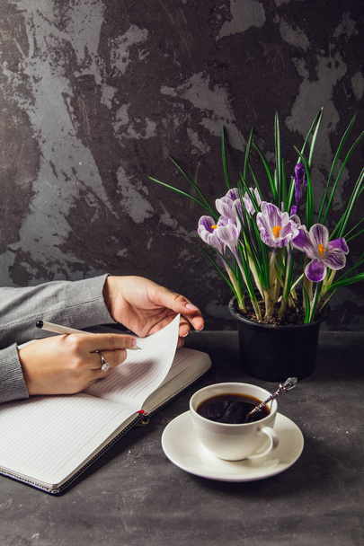 The girl's hands closeup with a pen in hand writing in a notebook. On the table is a Cup of coffee and a vase with flowers crocuses - Photo, Image