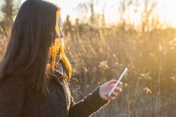 Chica joven sosteniendo el teléfono móvil en la luz del atardecer al aire libre
 - Foto, imagen