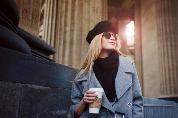 Young, hip and attractive blonde walking around the city with coffee to go, girl in a stylish hat and a gray coat - Foto, Imagen