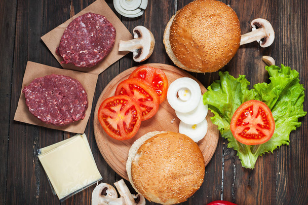 Homemade hamburger ingredients. Raw minced beef, fresh bun, slice of cheese, tomato, onion rings, lettuce on wood background - Photo, Image