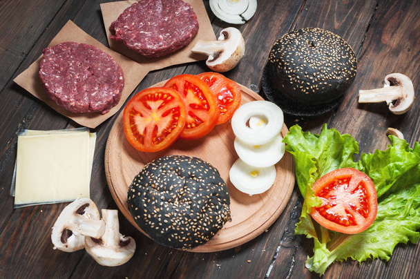 Homemade hamburger ingredients. Raw minced beef, fresh black bun, slice of cheese, tomato, onion rings, lettuce on wood background - Foto, afbeelding