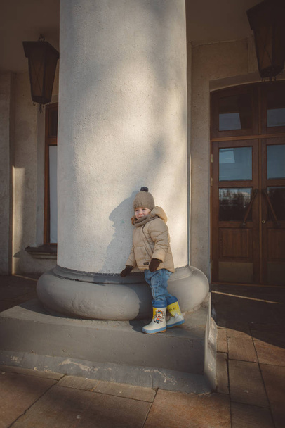 little girl in a beige jacket, jeans and rubber boots in the white columns, the entrance to the theater early spring, sunny day - Foto, Imagem