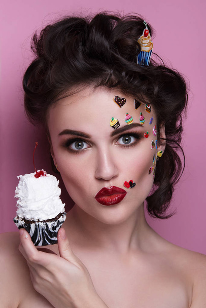 Beautiful fashion girl with luxury professional makeup and funny emoji stickers glued on the face. Young woman with cupcake in hands. - Foto, immagini