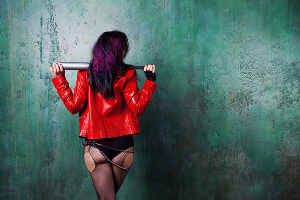 Punk woman with a bat in red leather jacket, standing against the wall, back. Free space on the right - Foto, Imagem