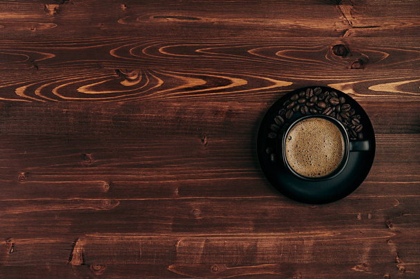 Hot coffee in black cup with crema and beans with copy space on brown old wooden board background, top view.  Rustic style. - Fotoğraf, Görsel