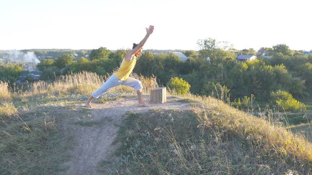 Joven deportista de pie en yoga posan al aire libre. Hombre caucásico practicando movimientos de yoga y posiciones en la naturaleza. Atleta balanceándose. Hermoso paisaje como fondo. Vida activa saludable
 - Foto, imagen