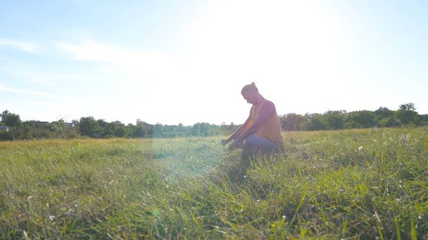 Nuori mies istuu vihreällä ruoholla niityllä ja harrastaa joogaa. Lihaksikas mies venyttelee ruumistaan luonnossa. Treenaa ulkona aurinkoisena päivänä. Kaunis maisema taustalla. Sulje se.
 - Valokuva, kuva