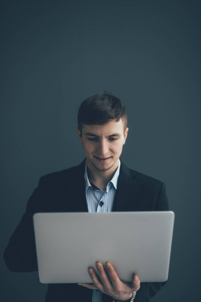 Knappe jonge zakenman is met behulp van een laptop, camera kijken en glimlachend, op zwarte achtergrond - Foto, afbeelding