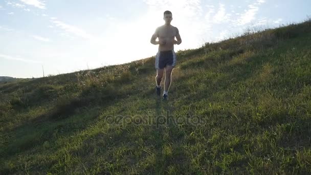 若い男は青い空を背景に緑の丘の上を実行しています。ジョギング男性アスリートは、日没で自然の中。スポーツ ランナー ジョギング フレアと日の出時の屋外の上り坂。クロスカントリー スキーのトレーニング。スローモーション - 映像、動画