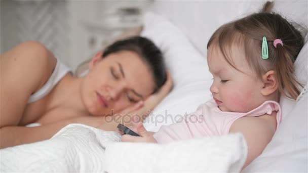 Closeup Portrait of Little Girl and Tired Mother in the Bedroom. Woman is Sleeping, Her Clever Daughter is Using Mothers Smartphone. - Materiaali, video
