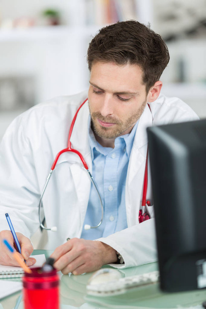attractive young doctor writing prescription sitting at desk - Foto, afbeelding