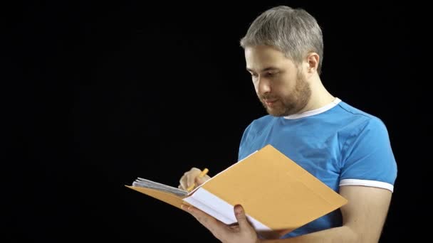 Handsome man in blue tshirt looking through papers in yellow folder. Cotract, bills, checklist concepts. Black background. 4K video - Video, Çekim
