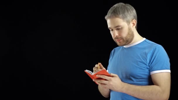 Handsome man in blue tshirt writing in his red notebook. Memo, search or contact concepts. Black background. 4K video - Video