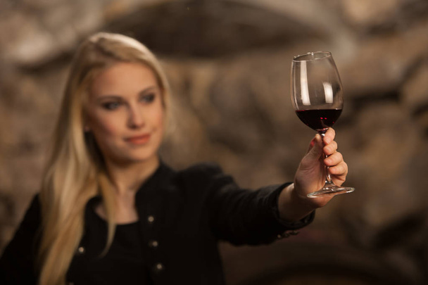 Beautiful young blond woman drinks wine in wine cellar - Фото, изображение